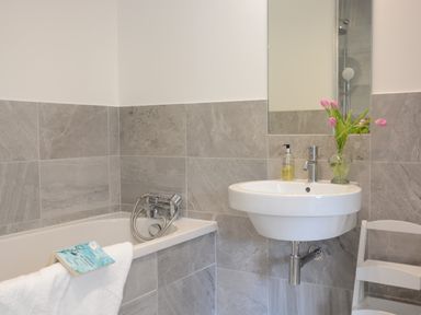 A bathroom with a bathtub a sink with a mirror a soap dispenser pink flowers in a vase and a white chair at 2 Pink Cottages Aldeburgh