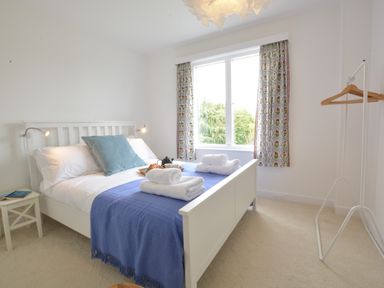 A bedroom with a white bed with blue blanket and folded towels near a window with patterned curtains at 2 Pink Cottages Aldeburgh
