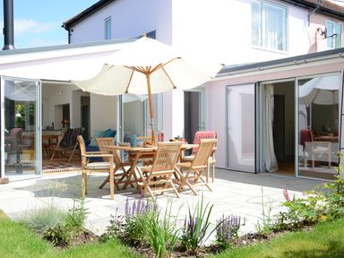 A patio with a wooden table and chairs under an umbrella outside a house with open glass doors at 2 Pink Cottages Aldeburgh
