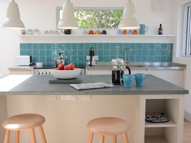 A kitchen with teal tiled backsplash a countertop with a bowl of apples French press coffee two blue mugs and wooden stools at 2 Pink Cottages Aldeburgh