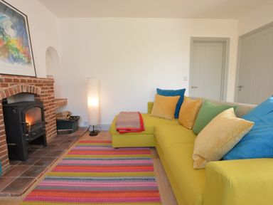 A living room with a yellow sofa and colorful cushions near a brick fireplace with a wood-burning stove at 2 Pink Cottages Aldeburgh