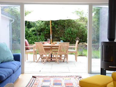 A patio with wooden table and chairs under a white umbrella viewed from a living room with blue sofa yellow chair and patterned rug at 2 Pink Cottages Aldeburgh