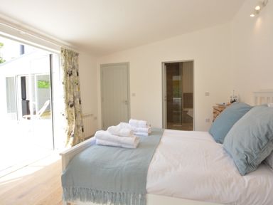 A bedroom with a white bed with blue pillows and blanket towels on bed a chest of drawers a door leading to a bathroom and patio doors at 2 Pink Cottages Aldeburgh