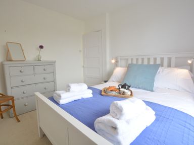 A bedroom with a white bed with blue bedspread and folded white towels on the bed a grey chest of drawers with a mirror and a flower vase on top and a wooden chair at 2 Pink Cottages Aldeburgh