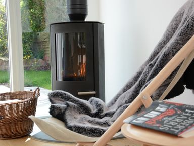 A modern black stove with fire burning near a wicker basket and a chair with a fur throw and a book on a wooden table at 2 Pink Cottages Aldeburgh
