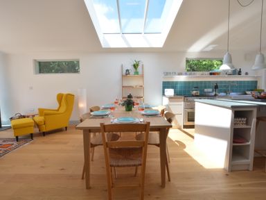 A dining area with a wooden table set for six and a yellow armchair near the kitchen at 2 Pink Cottages Aldeburgh
