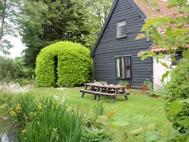 A garden with a green hedge, a wooden picnic table, and a black wooden house at Wassicks Cottage Haughley