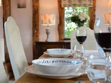 A dining table set with bowls plates wine glasses and water glasses in a room with wooden beams and a lamp at Wassicks Cottage Haughley