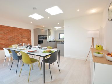 A dining area with a table set with plates glasses and bread surrounded by colorful chairs next to a kitchen with gray cabinets and a wooden sideboard with bowls of fruit at Skylight Aldeburgh in Aldeburgh