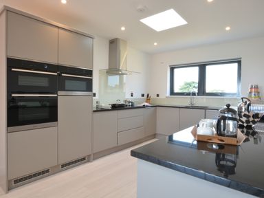 A modern kitchen with built-in ovens a stovetop under a glass hood a sink below a window and a countertop with a tray holding a teapot and cups at Skylight Aldeburgh in Aldeburgh