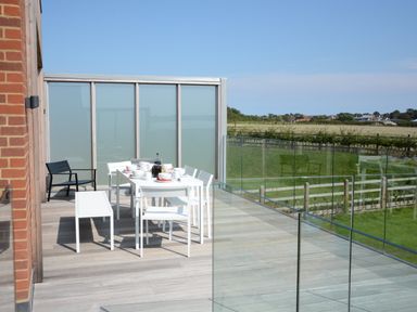 An outdoor terrace with a white dining table set with cups and plates and white chairs and benches with glass railing and a field view at Skylight Aldeburgh in Aldeburgh