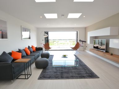 A living room with a dark sofa with orange and grey cushions a glass coffee table on a dark rug two brown chairs a TV on a wall-mounted wooden unit and a telescope by an open sliding door at Skylight Aldeburgh in Aldeburgh