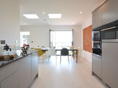 A kitchen with gray cabinets, a dining table with mixed color chairs and an open door to outside at Skylight Aldeburgh in Aldeburgh