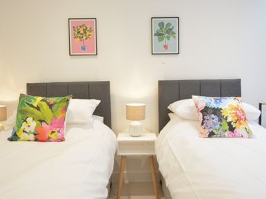 Two single beds with gray headboards and colorful pillows with a white nightstand and lamp between them with framed plant pictures on the wall at Skylight Aldeburgh in Aldeburgh