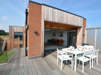 An outdoor dining table with white chairs on a wooden deck outside a room with folding glass doors at Skylight Aldeburgh in Aldeburgh