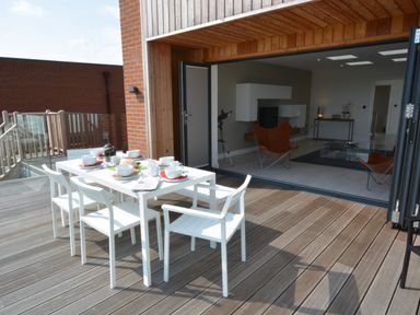 A dining table with white chairs set for a meal on a wooden deck outside a living room with orange chairs and a glass coffee table at Skylight Aldeburgh in Aldeburgh