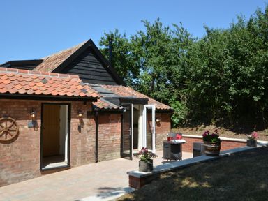 A brick house with tiled roof and open doors leading to a patio with a small table and two chairs with flowers on the wall at Carriage House Bruisyard Near Framlingham