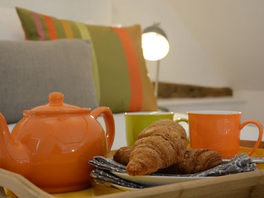 A breakfast tray with an orange teapot orange and green mugs croissants and pillows in the background at Carriage House Bruisyard Near Framlingham