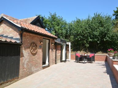 A brick patio with table and chairs with red cushions outside a building with tiled roof and wooden door at Carriage House Bruisyard Near Framlingham