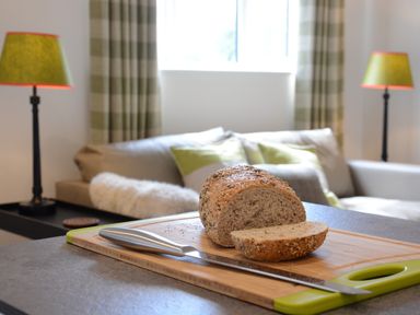 A kitchen counter with a cutting board holding a partially sliced loaf of seeded bread and a knife with a living room and two lamps in the background at Carriage House Bruisyard Near Framlingham
