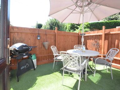 An outdoor patio area with a white table and chairs under a large umbrella and a barbecue grill against a wooden fence at Pine Cottage in Woodbridge