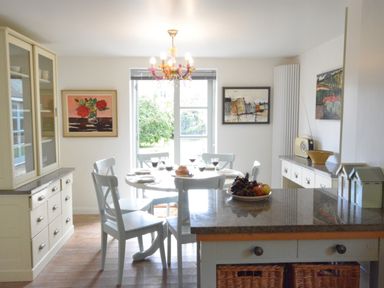 A dining room with a round table set with plates and wine glasses surrounded by chairs and a kitchen island with a bowl of fruit at Robert Sadler's Studio in Aldeburgh