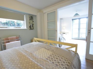 A bedroom with a patterned bedspread and towels on a rack next to a window and a view into a bright adjoining room at Robert Sadler's Studio in Aldeburgh