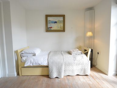 A bedroom with a single bed covered with white bedding and a throw blanket with folded towels on top a bedside table with a lamp and a painting on the wall at Robert Sadler's Studio in Aldeburgh