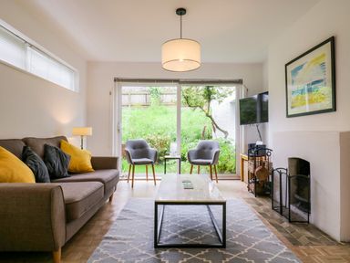 A living room with a sofa with yellow and dark pillows two gray chairs a coffee table a fireplace and a wall-mounted TV at The Bridge House in Southwold
