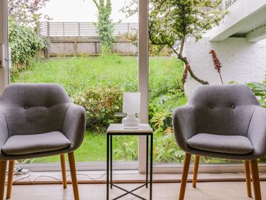 A sitting area with two grey chairs and a small table with a white lamp in front of a glass window showing a garden at The Bridge House Southwold