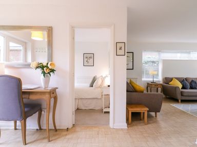 A bedroom with a bed and framed pictures viewed through a doorway from a living area with sofas and cushions and a desk with flowers at The Bridge House Southwold