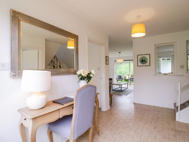 A hallway with a wooden desk chair and lamp next to a large mirror and leading to a living room with chairs and a coffee table at The Bridge House Southwold