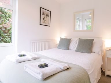 A bedroom with a bed with green pillows and white linens with towels on the bed a bedside table with a lamp a window with greenery outside a framed picture on the wall and a mirror at The Bridge House in Southwold