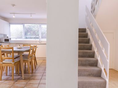 A kitchen with wooden dining table and chairs next to carpeted stairs with white railing at The Bridge House Southwold