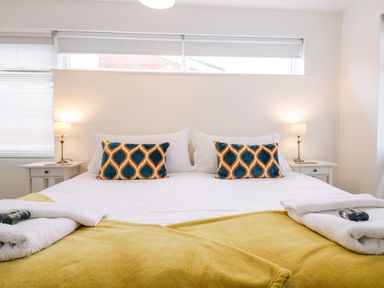 A bedroom with a large bed two patterned pillows yellow bed cover white towels on bed and bedside tables with lamps at The Bridge House Southwold