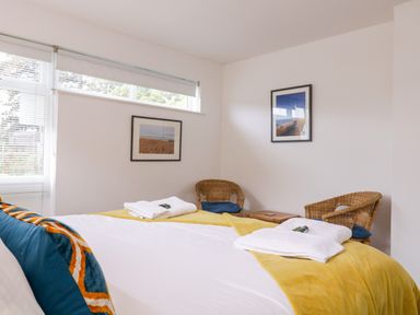 A bedroom with a bed covered by white and yellow bedding two wicker chairs with blue cushions and framed pictures on the walls at The Bridge House Southwold