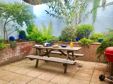 An outdoor patio with a wooden picnic table set with plates and a bottle of juice surrounded by brick planter walls with green plants and a red barbecue at The Red Brick House Aldeburgh