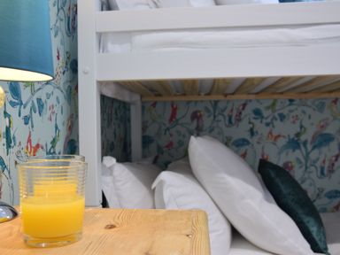 A bedside table with a glass of orange juice next to a bunk bed with pillows and bird patterned wallpaper at The Red Brick House Aldeburgh