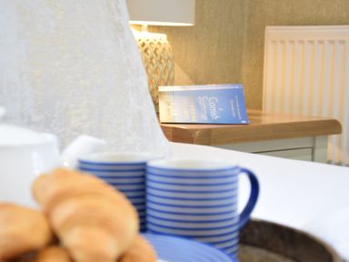 A bedside table with a blue book a lamp and part of a tea set with croissants on a bed at The Red Brick House in Aldeburgh