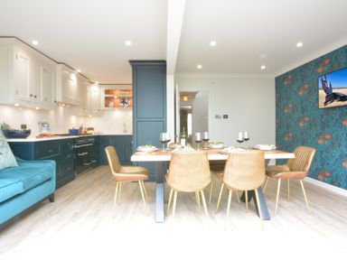 A kitchen and dining area with a wooden table set with plates and wine glasses surrounded by brown chairs with a teal couch to the side and a blue floral wallpaper at The Red Brick House in Aldeburgh