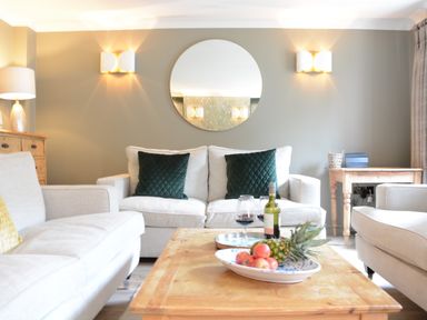 A living room with beige sofas green cushions a wooden coffee table with fruit and wine glasses a round wall mirror and wooden furniture at The Red Brick House Aldeburgh