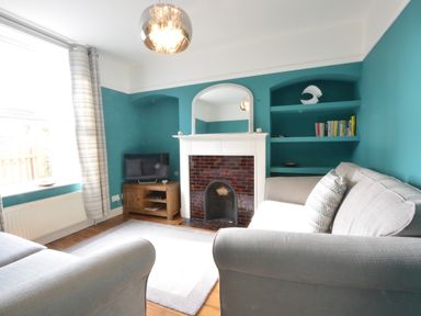 A living room with teal walls a fireplace a mirror a television on a wooden cabinet shelves with books and decorative items grey sofas and a window with patterned curtains at Somewhere Only We Know in Aldeburgh