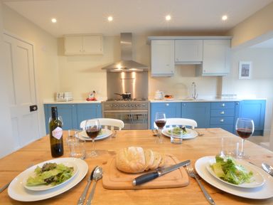 A kitchen with blue and white cabinets a dining table set with plates salad glasses and a loaf of bread at Somewhere Only We Know Aldeburgh