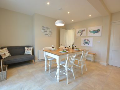 A dining area with a wooden table set for four people with white chairs and a gray sofa with cushions at Somewhere Only We Know in Aldeburgh