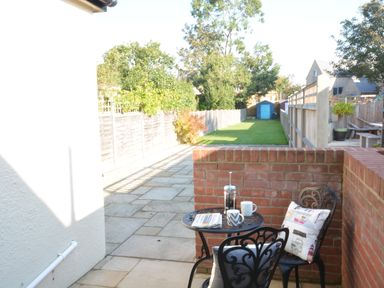 A small patio area with a table and two chairs next to a brick wall overlooking a long garden with a blue shed at Somewhere Only We Know Aldeburgh
