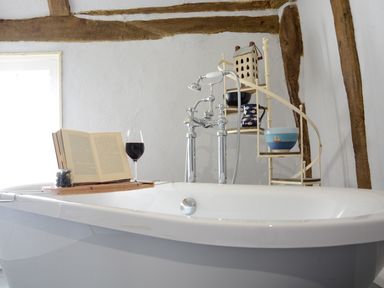 A bathroom with a white bathtub wooden beams on the wall a spiral shelf with bowls a book and a glass of wine on the bathtub at The White House with Studio in Wortwell