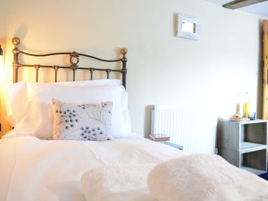A bedroom with a metal bed frame white bedding a floral pillow white towels a radiator a small table with books and a wooden shelf with juice and a croissant at The White House with Studio in Wortwell