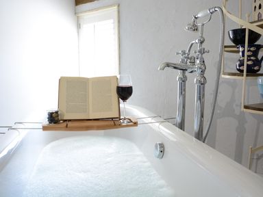A bathtub with foam water and a wooden tray holding an open book a glass of red wine and a jar of olives at The White House with Studio in Wortwell