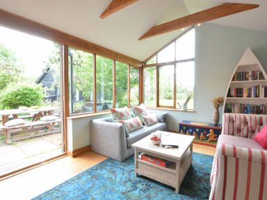 A living room with a gray sofa, striped armchair, blue rug, wooden coffee table, boat shaped bookshelf and windows overlooking a garden at The White House with Studio in Wortwell