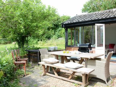 An outdoor patio area with a wooden table, benches, chairs, a barbecue grill, and greenery at The White House with Studio in Wortwell